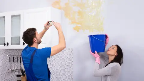 Person photographing a stained ceiling while another person holds a bucket under the drip